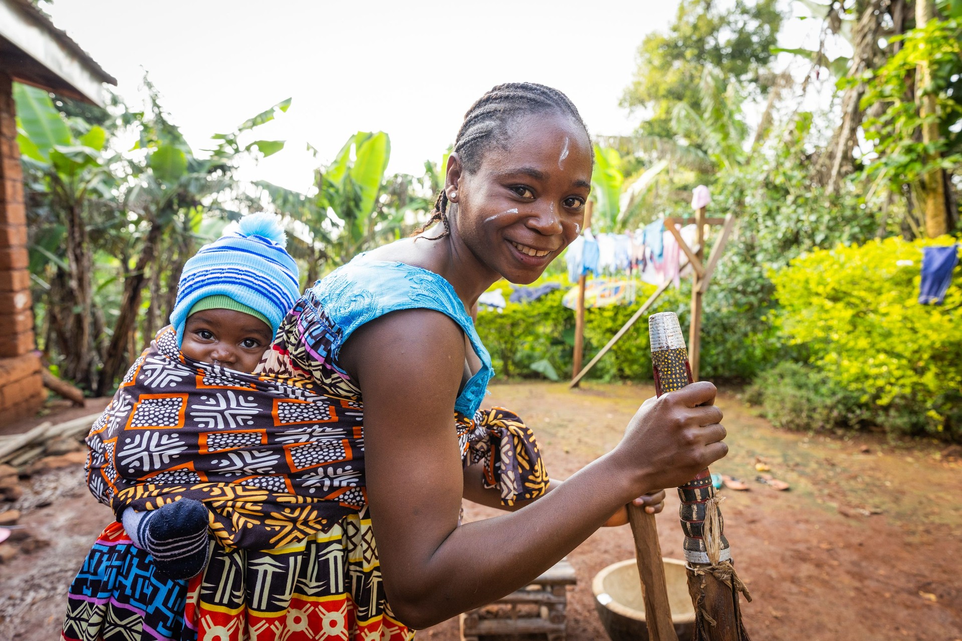 Outdoor scene featuring a woman carrying a child on her back, wrapped in vibrant traditional fabric, symbolizing maternal strength and cultural heritage. Captured in a rural Northern Ghana setting, the image highlights BongoAteel-Taaba’s capacity-building initiative for women, with laundry drying in the background and greenery surrounding the subject. Ideal for content focused on women’s empowerment, grassroots development, and community-led training programs in West Africa.